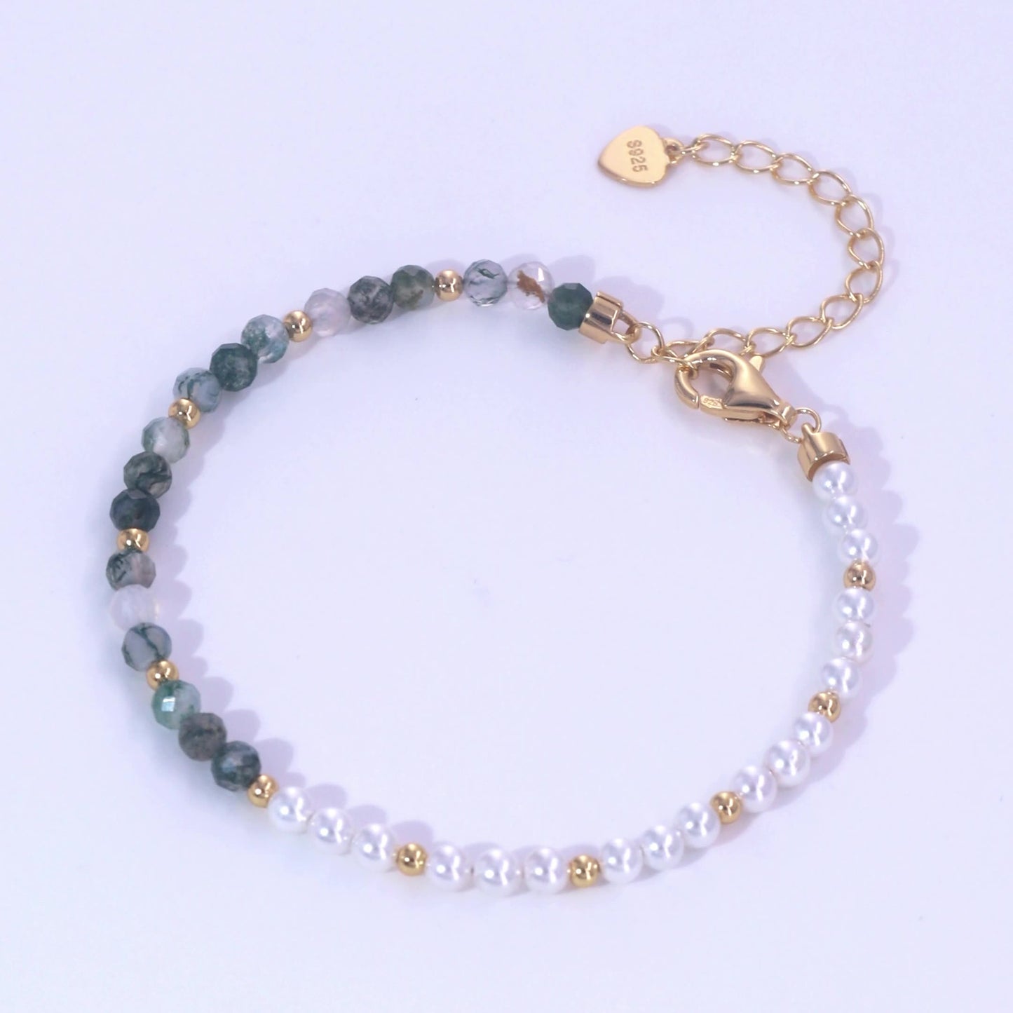 Gold - toned beaded bracelet: White pearls, moss agate stones, gold spacers, chain extender with “925” heart tag, light purple background.