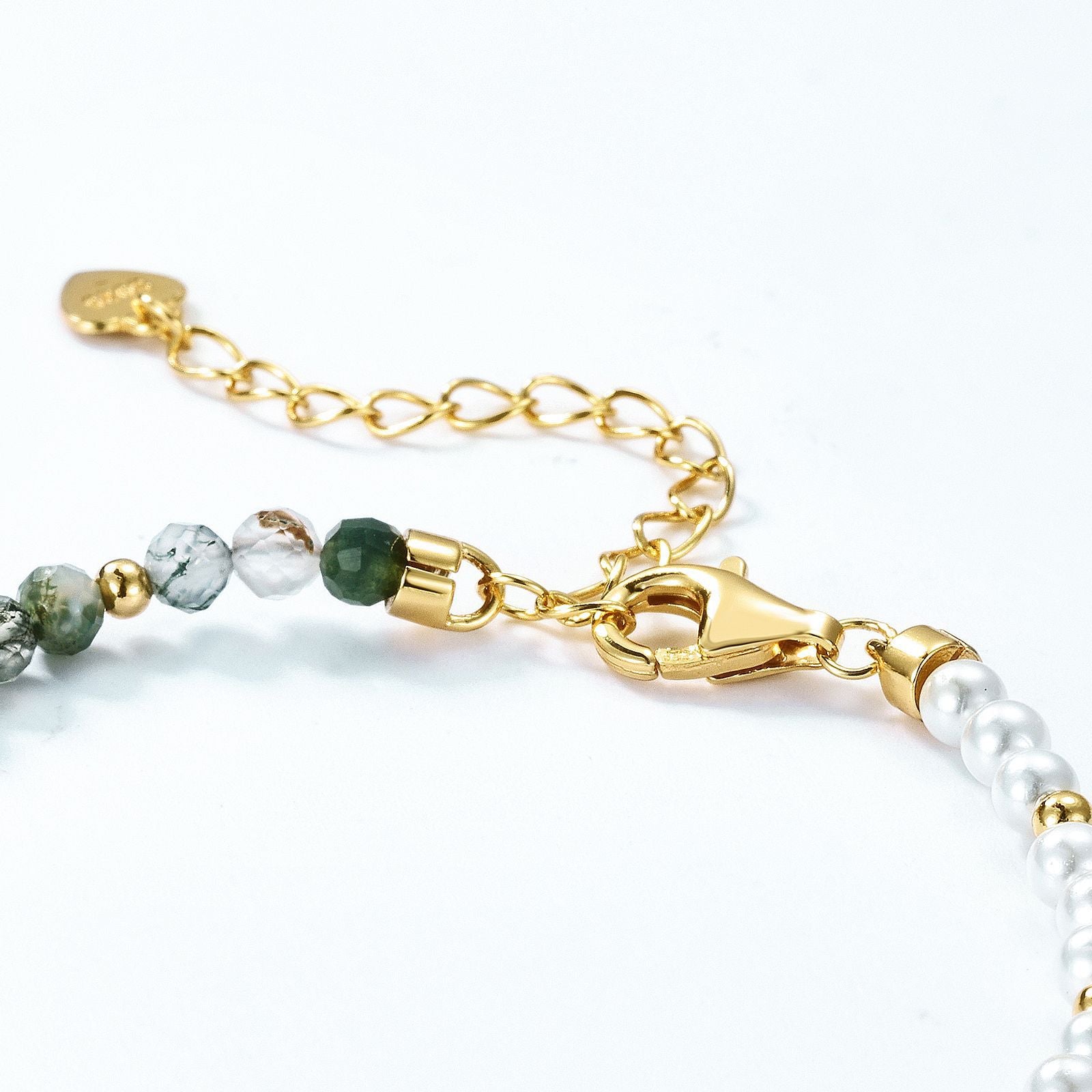 Close - up of gold - toned beaded bracelet clasp: Chain extender with heart tag, white pearls, green stones, gold spacers, white background.