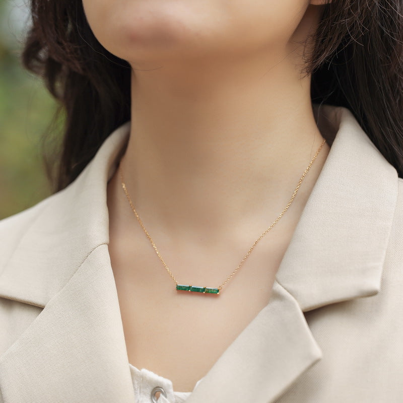Close - up of a person wearing gold - toned necklace: Horizontal bar pendant with three emerald - cut green stones, showcasing elegant style.