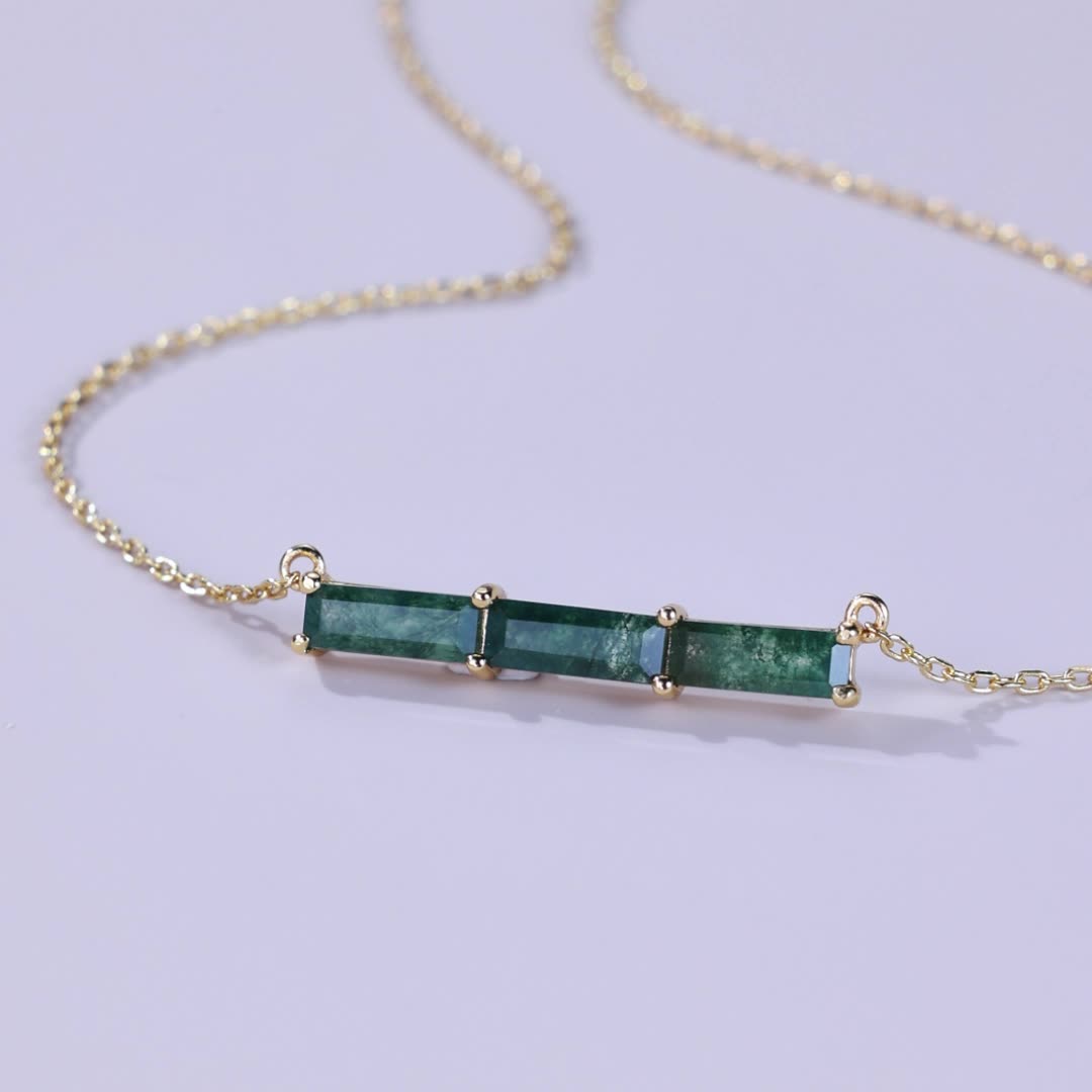 Gold - toned necklace: Horizontal bar pendant with three emerald - cut green stones, on a light purple background.