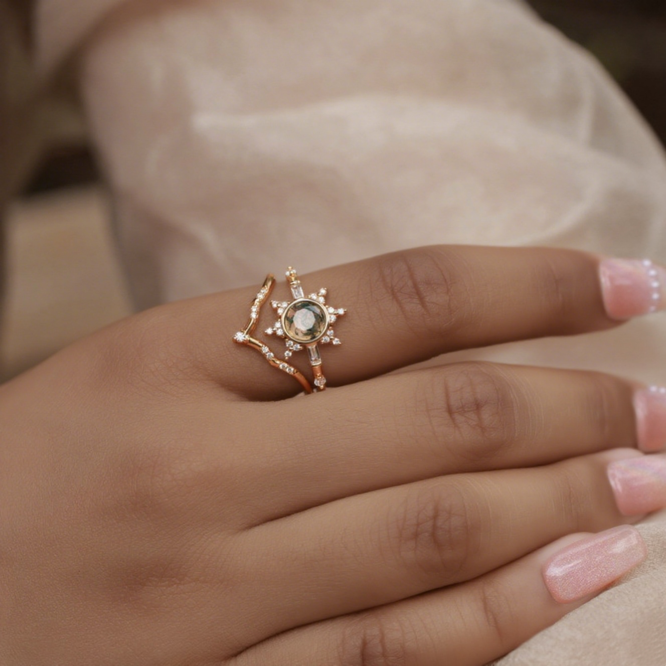 Gold - toned ring set with a sun - shaped moss agate ring and a curved crystal - studded ring, worn on a hand with light pink nails.