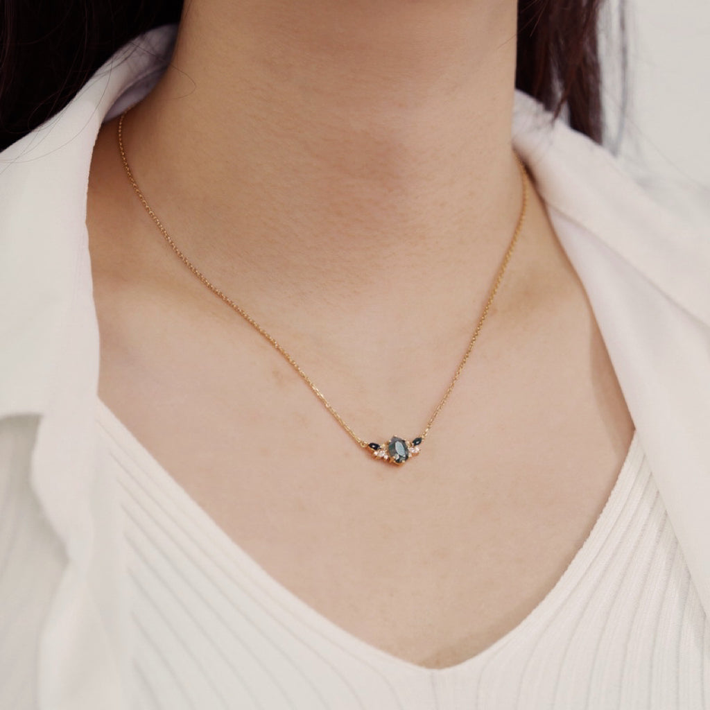 A person wearing a gold necklace: Features an oval moss agate pendant with green gemstone, opal, and crystal accents, paired with a white top.
