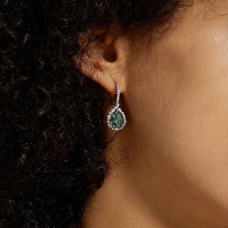Close - up of silver - toned earring on a person: Teardrop moss agate, crystal halo, hoop design, with curly hair.