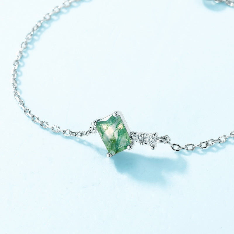 Close - up of silver bracelet charm: Rectangular moss agate, clear crystal accent, light blue background, highlighting detail.