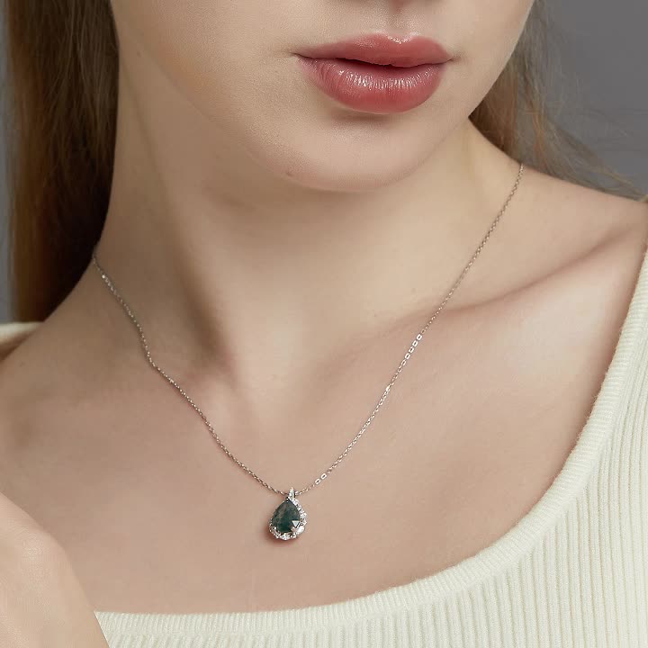 Person wearing silver pendant necklace: Teardrop - shaped moss agate pendant, light - colored top, gray background.