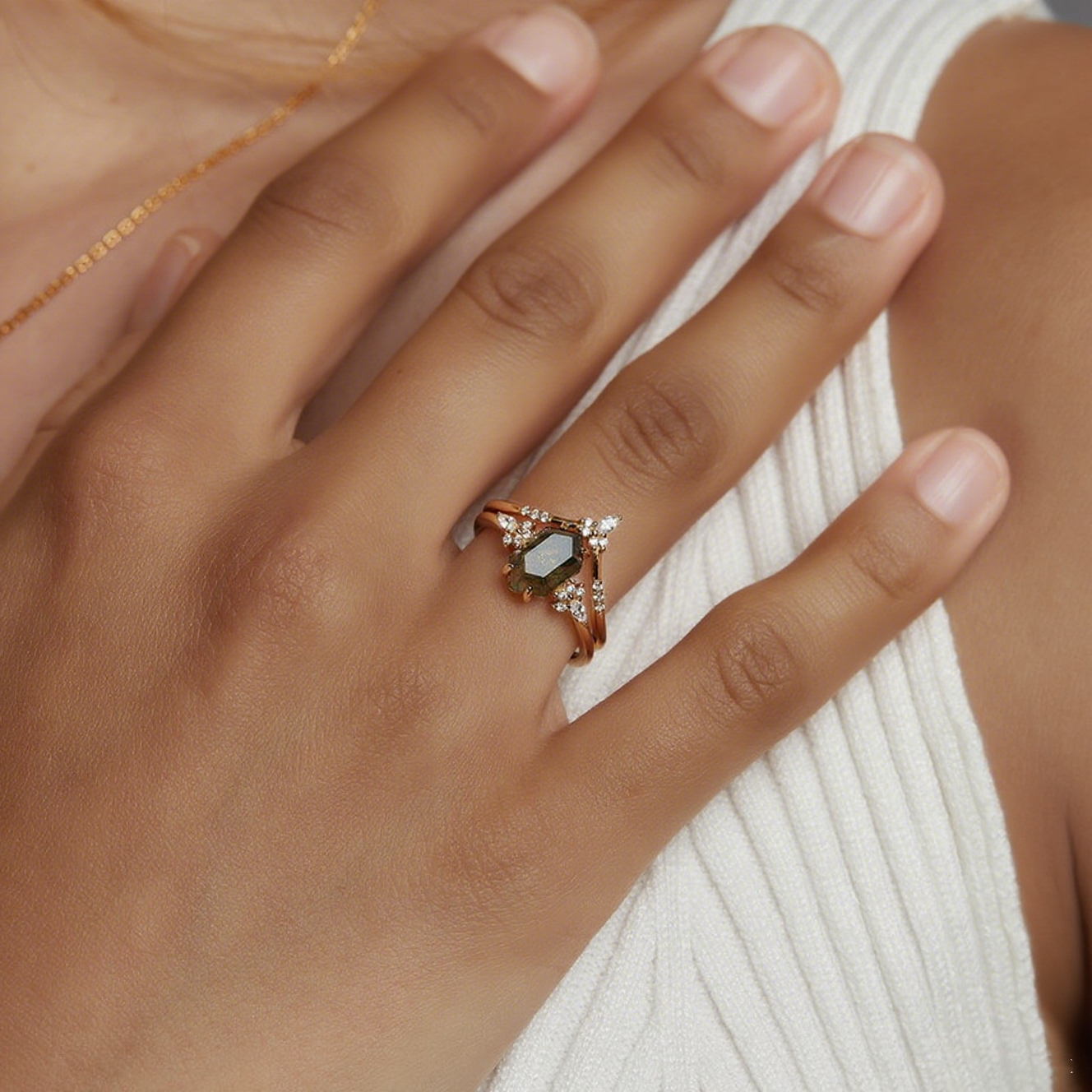 Hands wearing gold - toned ring set: Hexagonal moss agate ring with crystal accents, on a white fabric.