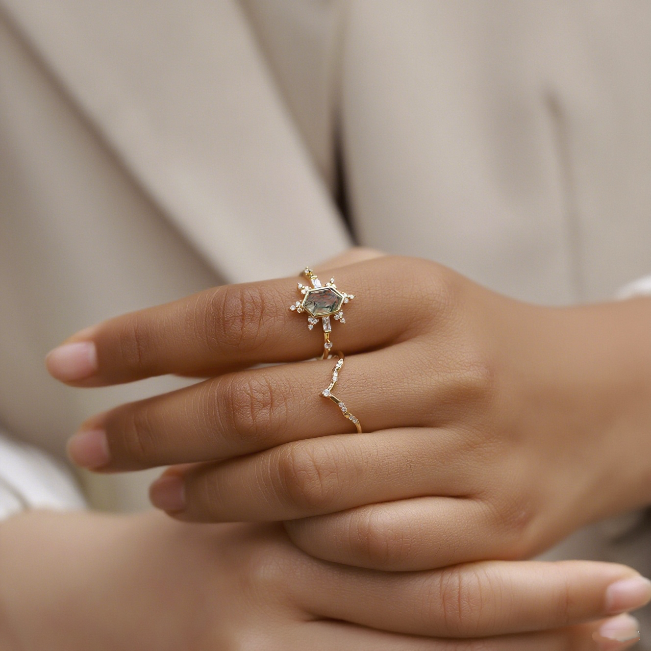 Hands wearing gold - toned ring set: Hexagonal moss agate ring and curved crystal - studded ring, on a light fabric.