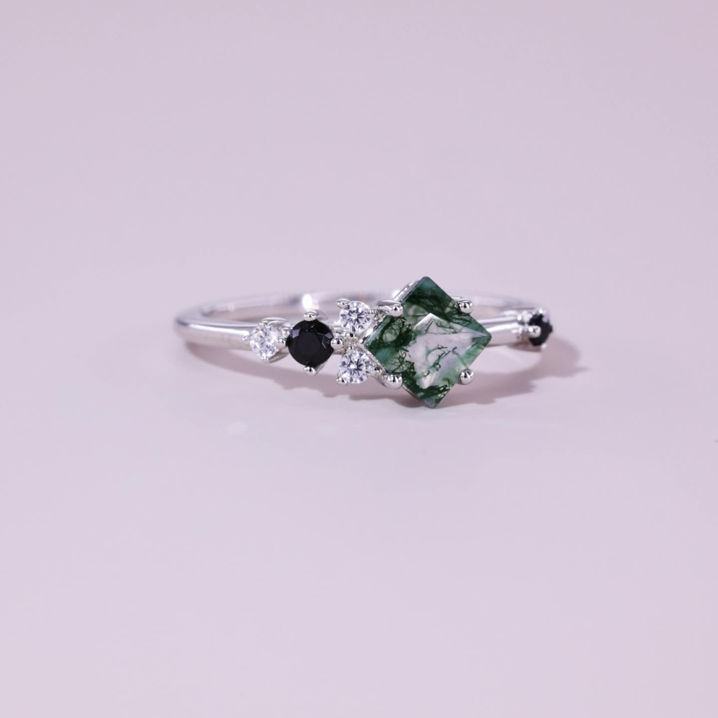 Silver ring with square - cut moss agate, flanked by black gemstones and clear crystals, set against a light pink background.
