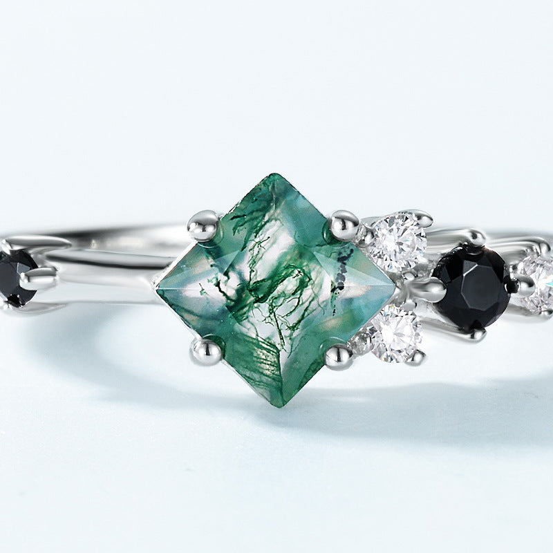 Extreme close - up of silver ring: Highlights the intricate patterns in the princess - cut moss agate, paired with black gemstones and clear crystals, on a light blue backdrop.