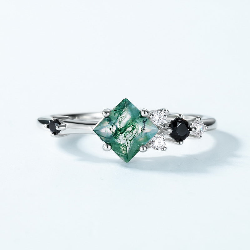 Silver ring: Square moss agate center, black gemstone and crystal - studded sides, presented on a light blue background, emphasizing geometric elegance.