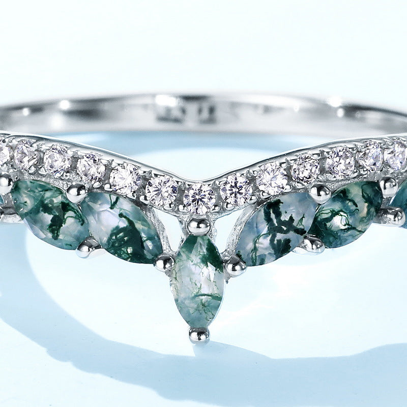 Silver ring featuring V - design, moss agate accents, close - up on light blue, emphasizing stone patterns.