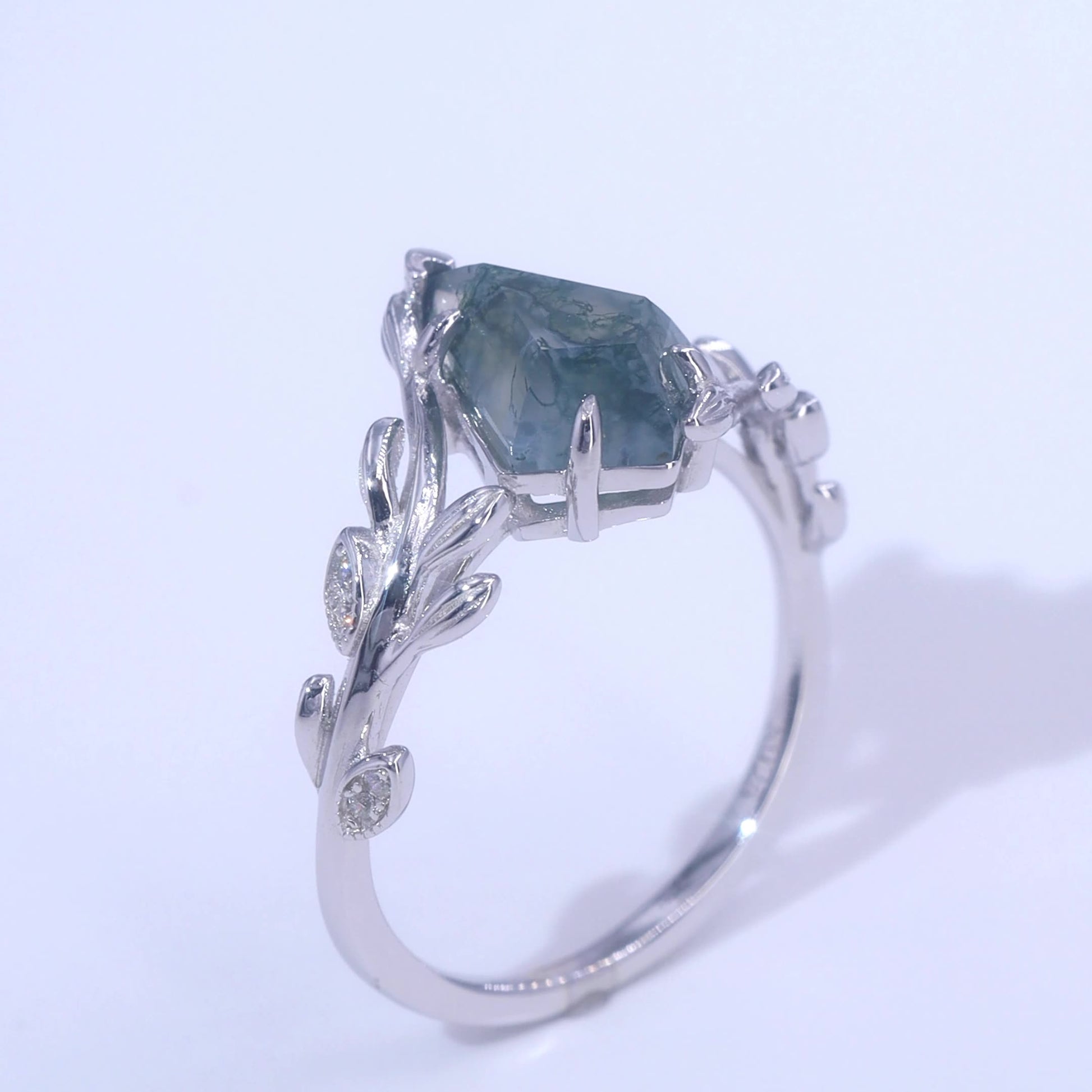Silver ring with a kite - shaped moss agate, adorned with leaf - branch motifs and small diamonds, set against a light pink background.