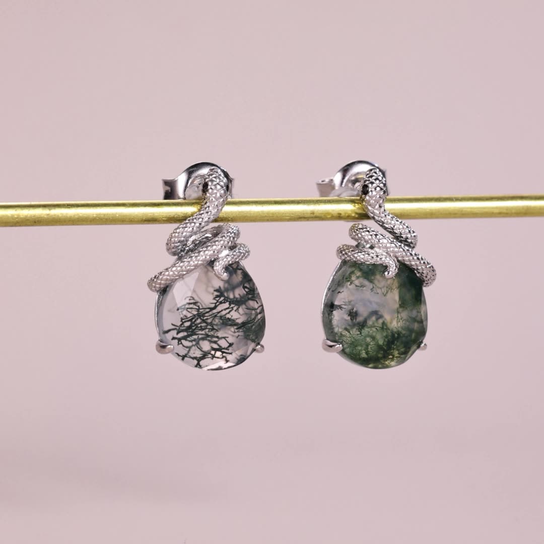 Silver - toned earrings: Snake - wrapped teardrop moss agate, hanging on a gold rod against a pink background.