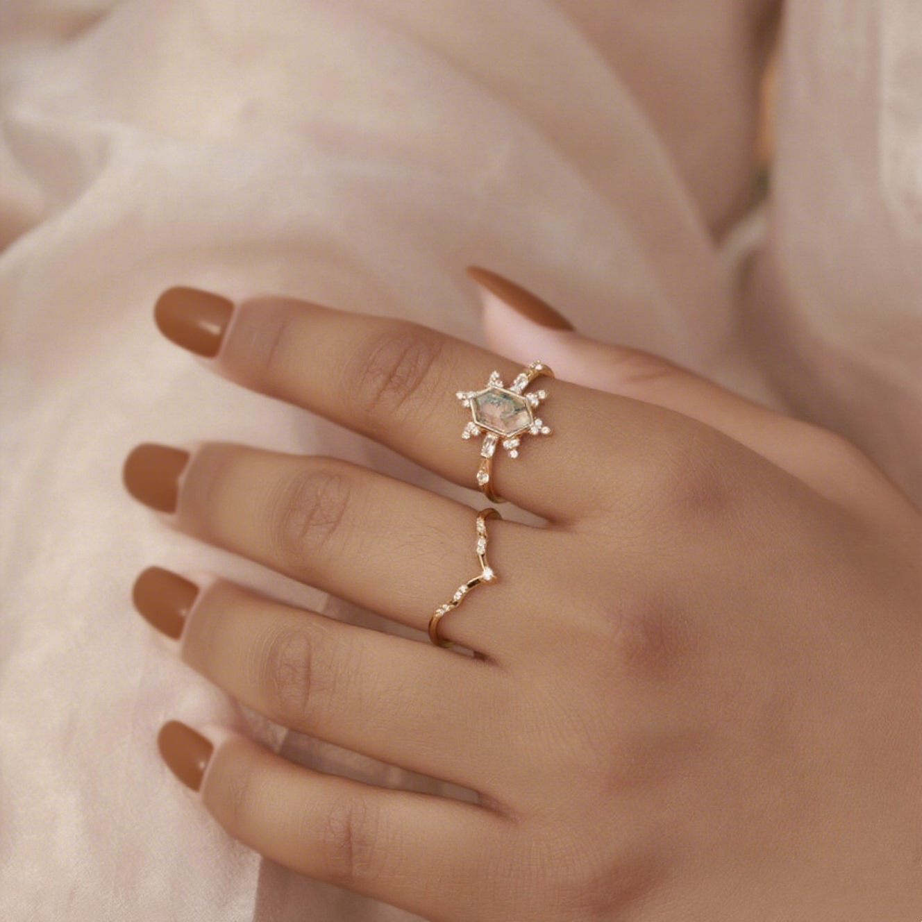 Hands wearing gold - toned ring set: Hexagonal moss agate ring and curved crystal - studded ring, on a light fabric.